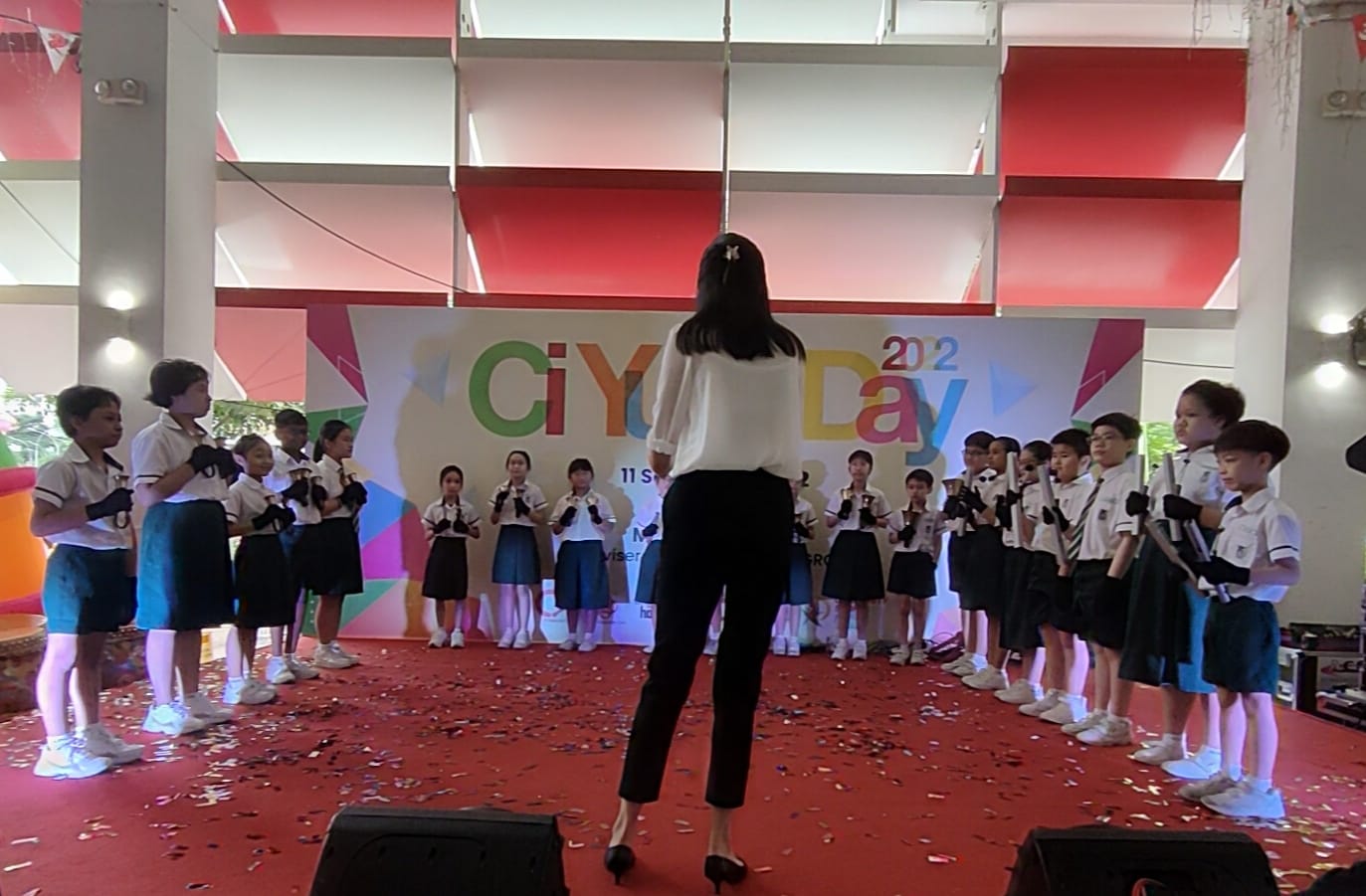 A woman in white top conducts uniformed schoolchildren performing on a red stage at CiYu Day 2022.